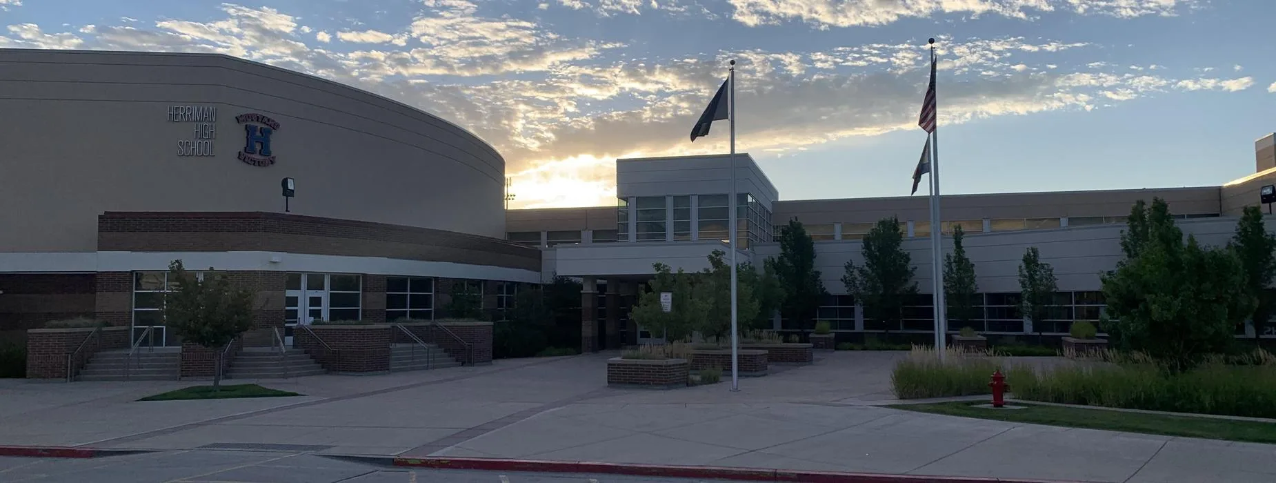 Herriman High School seen from the front car park at sunrise, the curved main entrance on the left and flagpoles on the right.